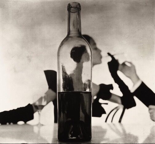 Irving Penn - Seattle Artist Black and white image of a woman photographed through the filter of a wine bottle in the foreground. The woman is in formal dress and opera gloves having her cigarette lit by a man arms coming from out of the frame. Photograph by Irving Penn once display in Capitol Hill Art Walk at Aria Salon in Seattle